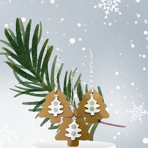 May include: Three brown paper Christmas tree ornaments with white glitter accents. The ornaments are hanging from twine and are against a light gray background with white snowflakes.