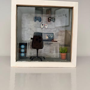 Puede incluir: Un diorama en miniatura dentro de una caja que muestra una configuración de juego con una silla negra, un monitor de computadora, una torre con luces circulares azules y una pequeña planta en maceta. La pared tiene un patrón de mármol.