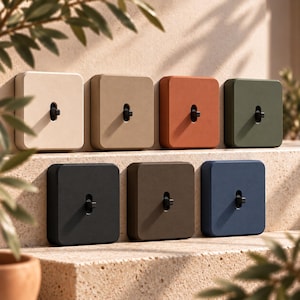 May include: A collection of seven square light switches with matte finishes in various colours including cream, tan, terracotta, olive green, black, brown, and navy blue. Each switch features a central black toggle lever.