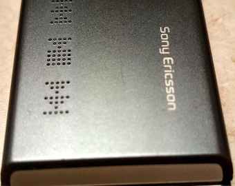Può includere: Un telefono cellulare a conchiglia Sony Ericsson Walkman nero con superficie testurizzata, obiettivo della fotocamera e icone di controllo multimediale. Il dispositivo mostra il marchio Sony Ericsson e il logo Walkman sulla cerniera argentata.