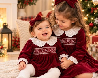 May include: Two matching long-sleeved dresses in deep red velvet, featuring white scalloped lace collars and cuffs with intricate white smocking detail across the chest. Each dress is paired with a large matching red velvet hair bow.