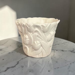 May include: A white ceramic planter with a detailed floral relief pattern. The small, round pot has a slightly flared rim and is suitable for small plants or decorative use. The planter is made of a matte white ceramic.