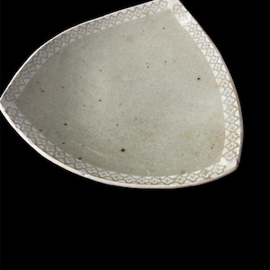 May include: A pale green, leaf-shaped ceramic plate with a decorative border. The border features a repeating pattern of small, stylized floral designs in a lighter shade of green. The plate has a slightly speckled surface.