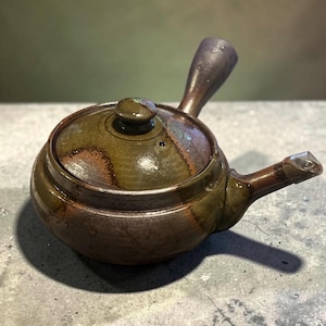 May include: A ceramic teapot with a side handle and a short spout, featuring a glossy glaze in shades of olive green, brown, and dark charcoal. The round body has a matching lid with a small knob, and the surface shows subtle color variations.
