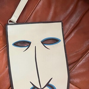Peut inclure: Un sac en forme de masque cr&egrave;me avec un contour noir et un design bleu clair et noir. Le sac a une longue et fine sangle cr&egrave;me attach&eacute;e &agrave; un anneau argent&eacute;. Le sac est sur une surface marron.