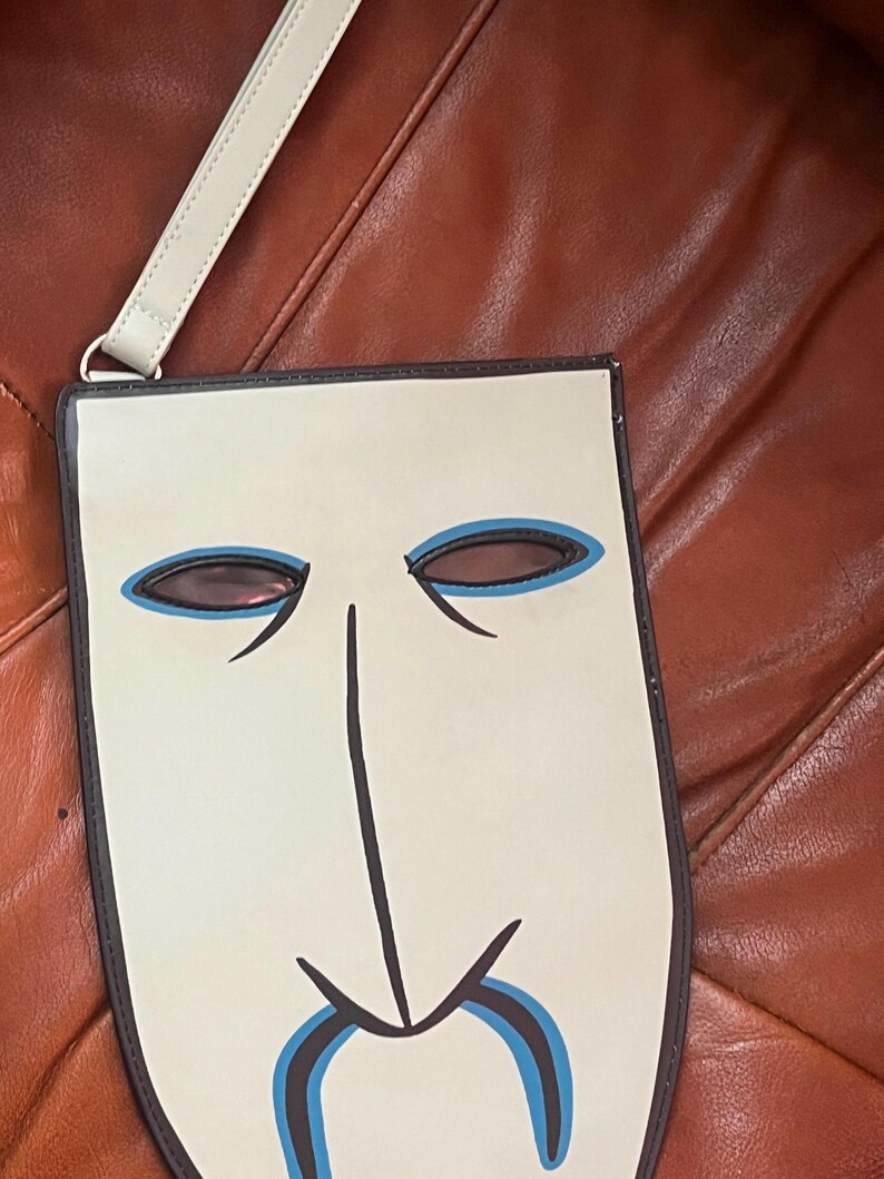 Peut inclure: Un sac en forme de masque cr&egrave;me avec un contour noir et un design bleu clair et noir. Le sac a une longue et fine sangle cr&egrave;me attach&eacute;e &agrave; un anneau argent&eacute;. Le sac est sur une surface marron.