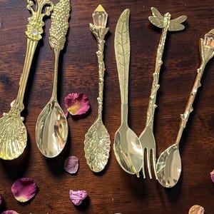 May include: An assortment of decorative gold-colored spoons and a fork with ornate designs. The spoons feature various handle embellishments, including a crystal, a leaf, and a dragonfly. Scattered rose petals add a touch of color.