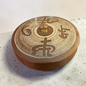 May include: A round, flat, brown ceramic lid with a central knob. The top surface features a light beige, circular glazed area decorated with dark brown, stylized calligraphic symbols arranged around the center knob, creating a distinct contrast.