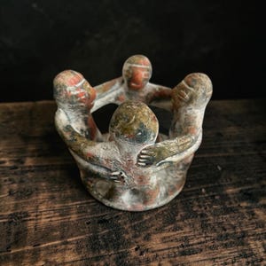 May include: A circular sculpture featuring four stylized figures standing in a ring, their arms linked around each other's shoulders. The piece is crafted from a mottled, textured material with earthy tones of reddish-brown, gray, and cream throughout.