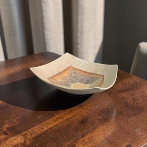 May include: A square ceramic bowl with curved edges, featuring a textured off-white rim and a central glazed area with blended tones of brown, gold, and grey. A small, embossed dragonfly motif is visible on one side of the textured border.