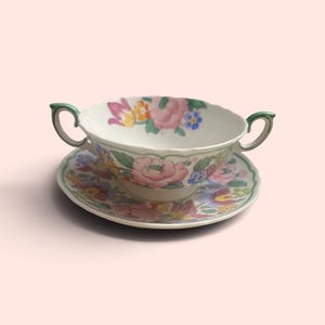 May include: A white porcelain bouillon cup and saucer set featuring a colorful floral pattern with pink, yellow, and blue flowers. The cup has two green-trimmed handles and a scalloped rim, while the saucer matches the floral design.