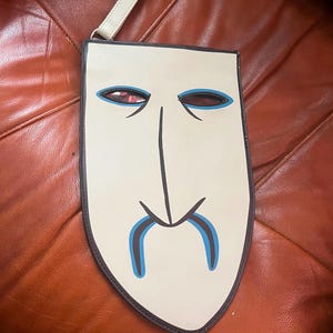 Peut inclure: Une pochette cr&egrave;me avec un motif de visage stylis&eacute;. Le visage pr&eacute;sente des lignes noires pour le nez et la bouche, avec des accents bleus autour des yeux et de la moustache. Les yeux ont un int&eacute;rieur rouge. Une petite sangle cr&egrave;me est attach&eacute;e.