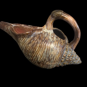 May include: A ceramic pitcher shaped like a conch shell with a glossy brown and cream glaze. The vessel features a textured surface with vertical ridges, a curved handle, and a wide spout. The base of the shell has a detailed, bumpy texture.