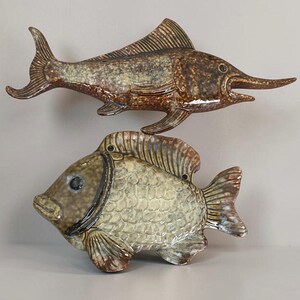 May include: Two ceramic fish wall decorations with a glossy glaze. The top fish is a long, slender marlin in shades of brown and tan. The bottom fish is a rounder, deep-bodied fish with a textured scale pattern in light beige and dark brown tones.