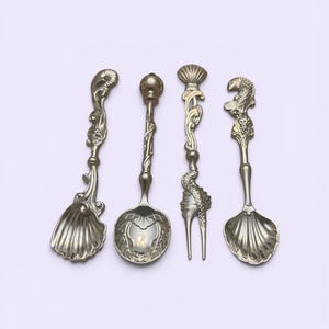 May include: Four ornate silver-colored serving utensils. The set includes two spoons, a fork, and a scoop, each with unique decorative handles and bowl designs. The handles feature seashell, floral, and aquatic creature motifs.