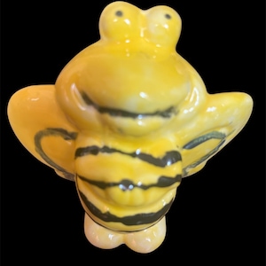 May include: A ceramic bee figurine with a glossy finish. The bee is primarily yellow with black stripes on its body and black detailing on its wings. The figurine has a whimsical, cartoonish design.