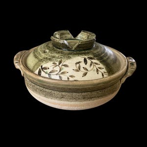May include: A round ceramic pot with a lid featuring a green and cream glaze. The lid is decorated with a dark brown leaf pattern and has a notched handle. The pot has two side handles and a textured, earthy finish with a band of green glaze.