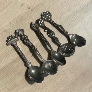 May include: A set of four silver-toned metal spoons with decorative handles. Each handle features distinct embossed designs including a shell, a mermaid, and intricate floral or geometric patterns. The spoon bowls vary in shape, including oval and shell-like forms.