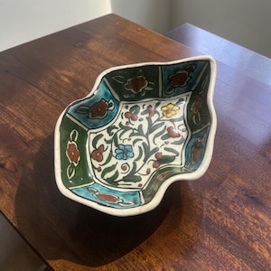 May include: A hand-painted ceramic bowl with an irregular, leaf-like shape. The interior features a central floral pattern with green, blue, yellow, and brown accents, surrounded by a border of alternating green and blue panels with floral motifs.