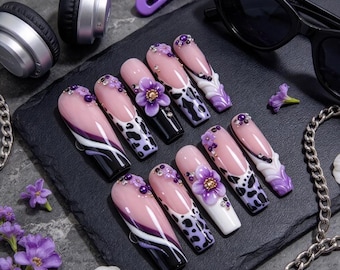 Pode incluir: Um conjunto de dez unhas postiças longas em formato de caixão com base nude, padrões de manchas de vaca em preto e roxo, espirais brancas e roxas, flores roxas em 3D e pequenos strass roxos dispostos sobre uma superfície escura.