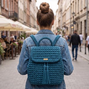 May include: A teal crochet backpack featuring a flap closure with a gold-toned rectangular brand tag and two matching hanging tassels. The bag has a top handle and two shoulder straps, all crafted from a thick, textured yarn material.