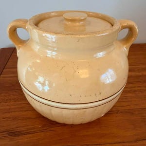 May include: A cream-colored ceramic crock with a matching lid and two side handles. The pot features a rounded body with a subtle horizontal brown line detail and a ribbed texture along the lower section, finished with a glossy glaze.