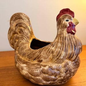 Può includere: Un vaso o ciotola in ceramica a forma di gallina con una smaltatura maculata marrone e crema. La figura presenta una cresta e bargigli rossi, un becco giallo e dettagliate texture di piume modellate sul corpo, sulle ali e sulla coda.
