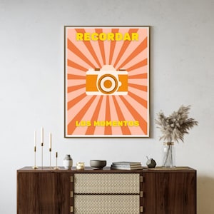 Könnte beinhalten: Ein rechteckiger Wandkunstdruck mit einem zentralen weißen und orangefarbenen Kamerasymbol vor einem Hintergrund aus abwechselnden hellrosa und orangefarbenen Sonnenstrahlen. Der gelbe Text "RECORDAR" steht über der Kamera und "LOS MOMENTOS" darunter.