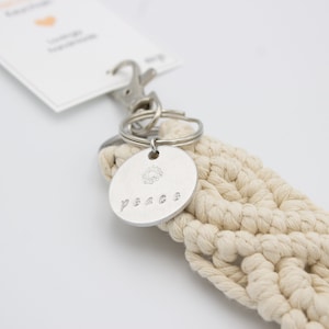May include: A macrame keychain with a silver-colored round charm. The charm has a small sunburst design and the word "peace" engraved on it. The keychain is attached to a metal clasp and a white tag.