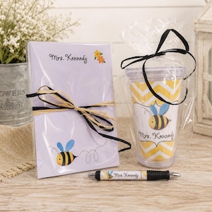 May include: A gift set featuring a notepad, a tumbler, and a pen, all with a bee theme. The notepad is tied with a black and tan ribbon and has the text "Mrs. Kennedy". The tumbler has a yellow and white chevron pattern and a bee graphic.