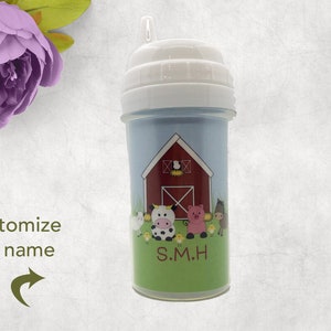 May include: A baby bottle with a white top and a blue and green body. The body features a red barn illustration with farm animals and the text "S.M.H." The bottle has a straw.