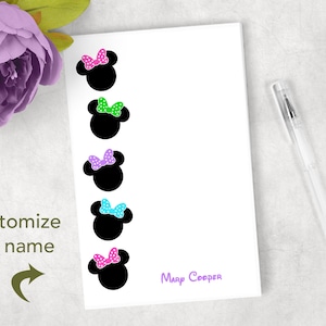 May include: A white notepad featuring black silhouettes of Minnie Mouse heads with colorful polka-dot bows in pink, green, purple, blue, and pink. The text "customize the name" and "Mary Cooper" are also visible. A clear pen rests on the side.