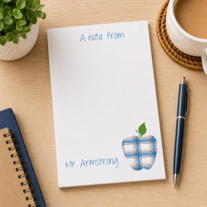 May include: A white rectangular notepad featuring the blue printed text A note from and Mr. Armstrong. The notepad includes a graphic of an apple with a blue and white plaid pattern and a green leaf. A dark blue pen rests beside the notepad.