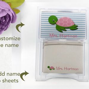 May include: A clear plastic dispenser with a stack of white notepads. The top notepad has a blue and white striped background with a pink and green turtle illustration and the text "Mrs. Hartman". The bottom notepad has a smaller turtle illustration and the text "Mrs. Hartman".