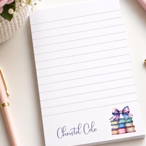 Op de afbeelding: Een wit rechthoekig notitieblok met horizontale lijnen. Het onderste gedeelte bevat de naam Christol Cole in blauw schrift, naast een aquarelillustratie van een stapel van drie boeken met een paarse en blauwe strik erbovenop.