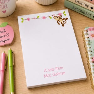 May include: White notepad with a monkey and floral design. The notepad has the text "A note from Mrs. Gelman" in pink. A pink and green pen are next to the notepad. A pink apple-shaped sticky note is also visible.