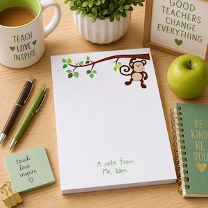 May include: A white notepad with a brown branch and monkey illustration. The notepad has the text "A note from Mr. Sam" in green. Other items include a mug, pens, a green apple, and a small notebook.