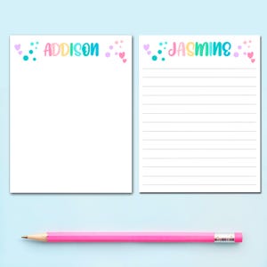 May include: Two white notepads with the names "Addison" and "Jasmine" in colorful letters. The "Addison" notepad is blank, while the "Jasmine" notepad has lined paper. A pink pencil with a white eraser rests below the notepads.