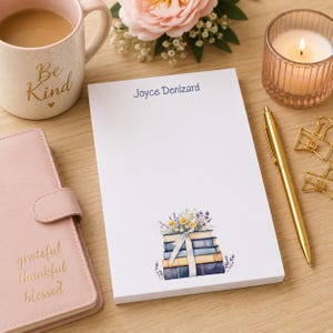 May include: A white notepad with the name "Joyce Denizard" printed in blue at the top. The bottom features a watercolor illustration of books topped with flowers and tied with a ribbon. A gold pen and paper clips are nearby.