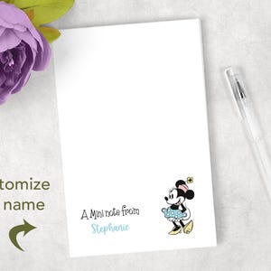 May include: A white notepad with a Minnie Mouse illustration and the text "A Mini note from Stephanie." A clear pen rests on the right. A purple flower is in the upper left corner. The words "customize the name" are on the left.