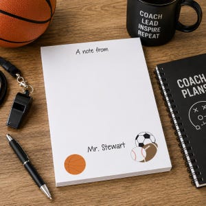 May include: A white notepad with the text "A note from" and "Mr. Stewart". The notepad has illustrations of a basketball, soccer ball, baseball, and football. A black pen, whistle, and mug are also visible.
