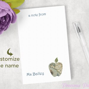 May include: White notepad with the text "a note from" and "Ms Bailey" in dark blue. An apple graphic with a map design is at the bottom. A clear pen rests on the right. A purple flower is in the upper left corner.