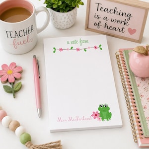 May include: A white notepad with a floral design and the text "a note from" and "Mrs. McFarland". A pink pen, a mug with "Teacher fuels" text, and a wooden flower are also visible. A small sign reads "Teaching is a work of heart".