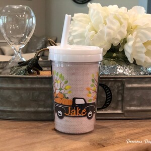 May include: A brown and tan reusable cup with a black truck and pumpkins on it. The truck has the name "Jake" written on the side. The cup has a straw and a black handle.