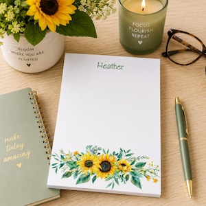 May include: A white notepad with the name "Heather" in green, and a sunflower and greenery design at the bottom. A green pen, a green notebook, a candle, and a small flower pot with a sunflower are also visible.