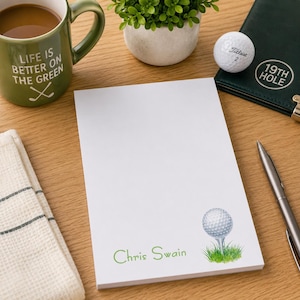 May include: A white notepad with a golf ball graphic and the name "Chris Swain" in green. A green mug with the text "Life is better on the green" and a golf club graphic is visible. A golf ball and a green notebook are also present.