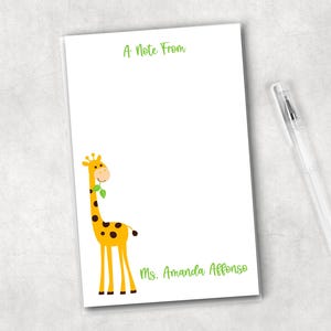 May include: White notepad with a cartoon giraffe illustration in yellow and brown. The notepad has the text "A Note From" at the top and "Ms. Amanda Affonso" at the bottom in green. A clear pen rests on the side.
