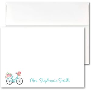 May include: White stationery set featuring a light blue bicycle with a basket of flowers. The bicycle is on the bottom left corner of the card. The text "Mrs. Stephanie Smith" is written in light blue script on the bottom right.