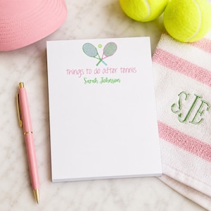 May include: A white notepad with the text "things to do after tennis" and a name below. The notepad has a tennis racket and ball graphic. A pink pen, tennis balls, a pink hat, and a striped towel with initials are also visible.