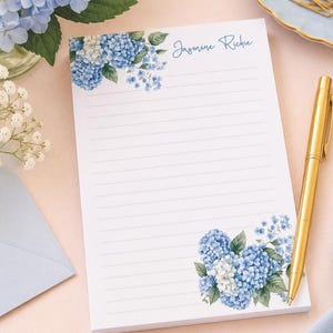 May include: A white rectangular notepad with horizontal lines, featuring blue hydrangea floral illustrations in the top left and bottom right corners. The name Jasmine Richie is printed in blue script at the top right of the page.